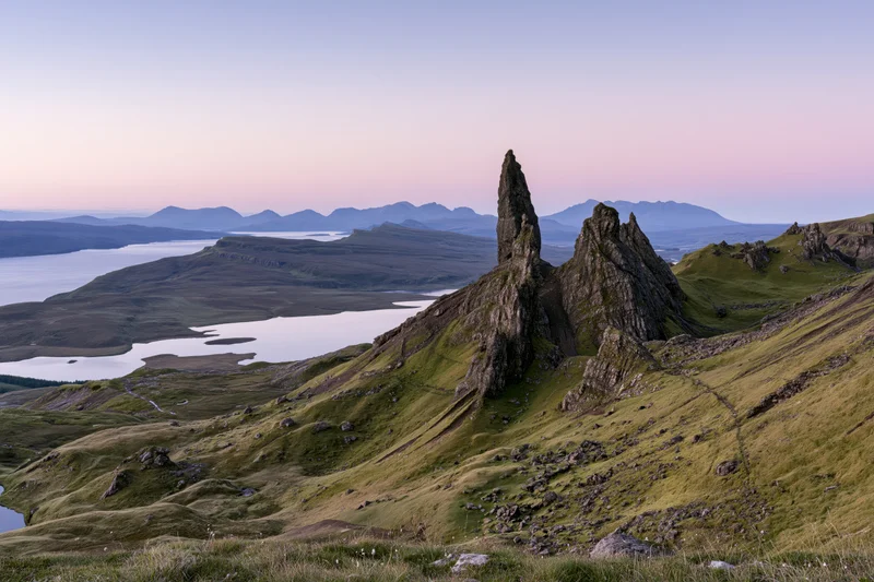 Old Man of Storr rock pinnacle rising from green hillside overlooking loch and distant mountains at twilight