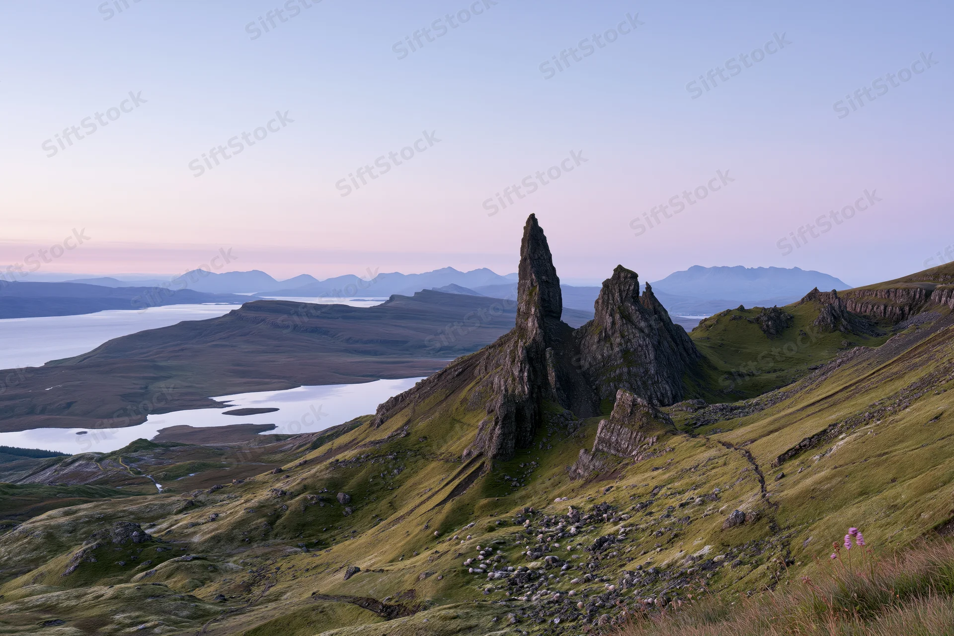Old Man of Storr rock pinnacle rising from green highland ridge overlooking loch and distant mountains at twilight