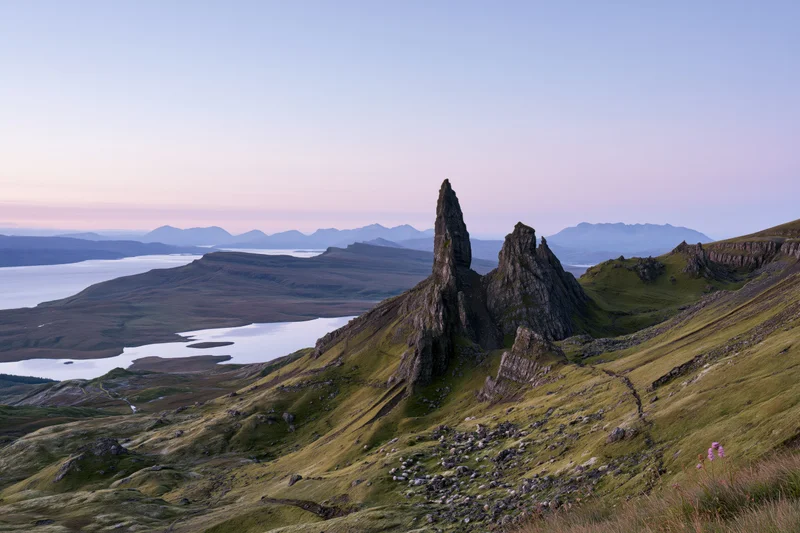 Old Man of Storr rock pinnacle rising from green highland ridge overlooking loch and distant mountains at twilight