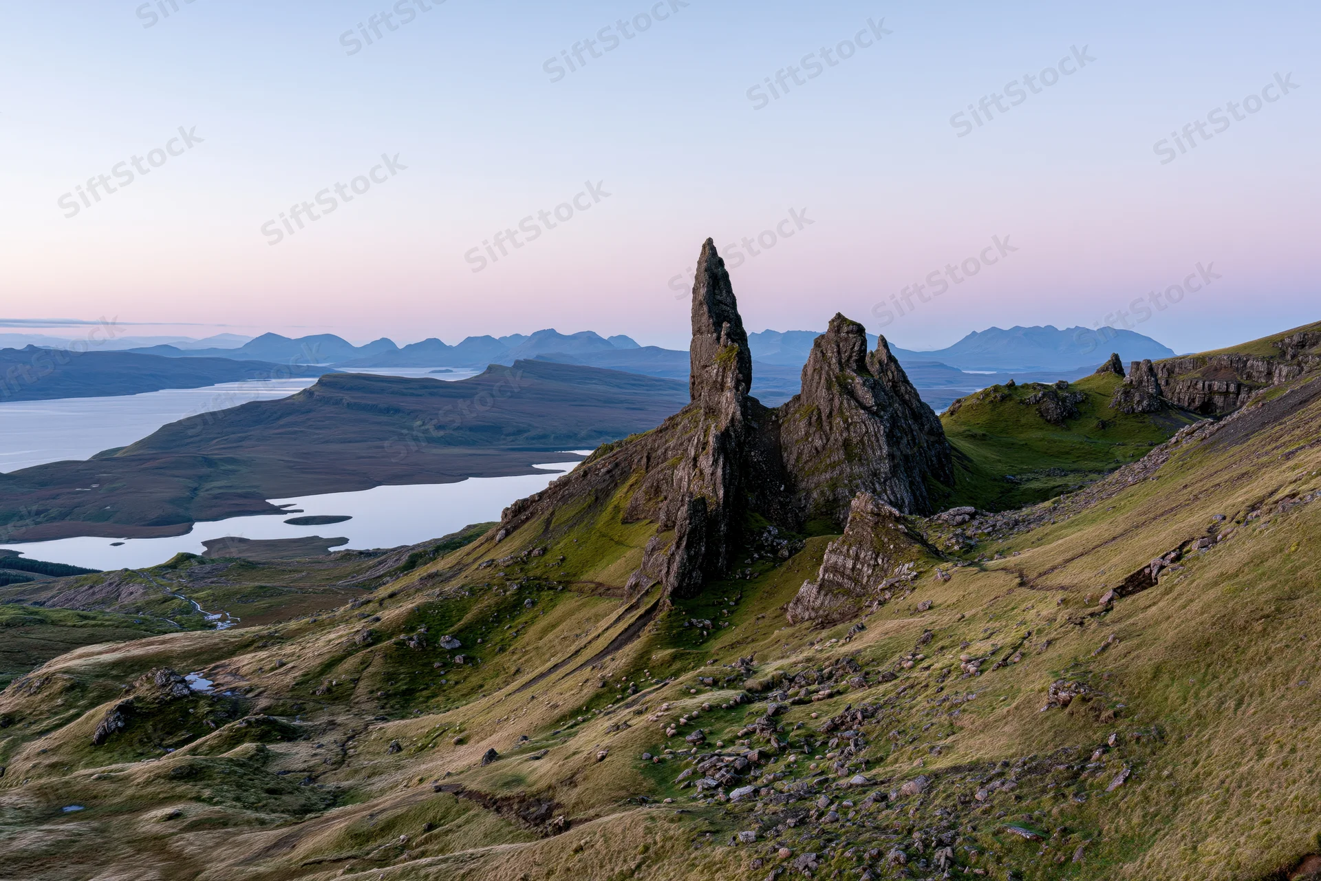 Rock pinnacle rising from grassy hillside overlooking loch and distant mountains at dusk