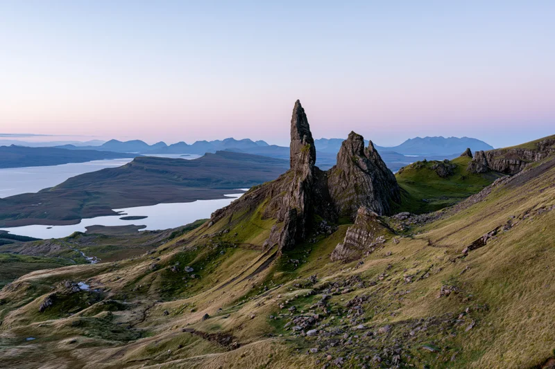 Rock pinnacle rising from grassy hillside overlooking loch and distant mountains at dusk