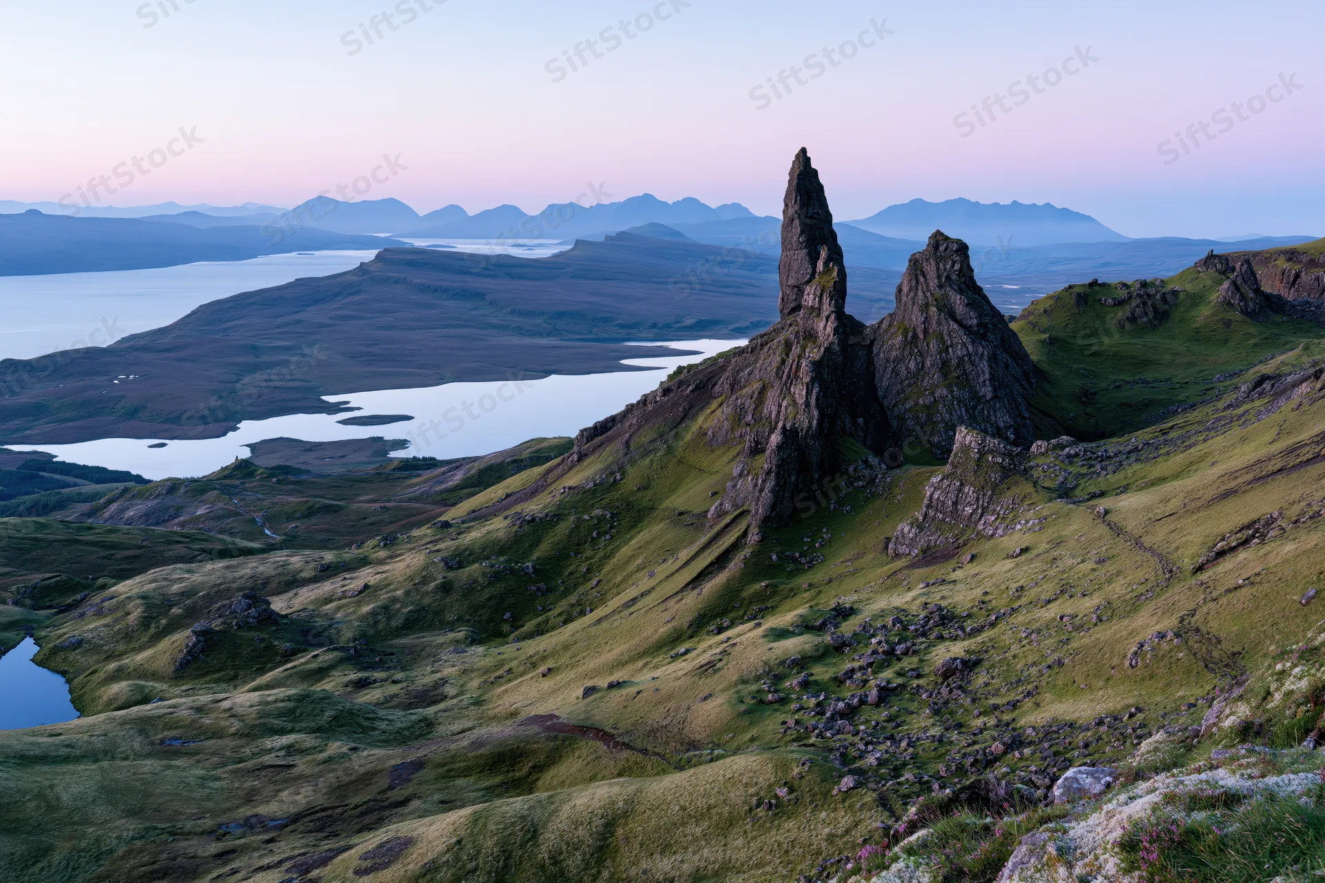 Old Man of Storr rock pinnacle rising from green highland hills overlooking lochs and distant mountains at twilight