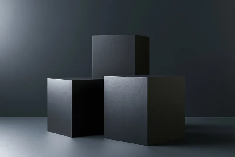 Three matte black geometric cubes arranged as pedestals in a dark studio space