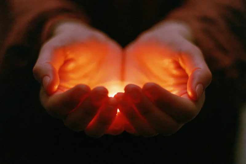 Cupped hands holding a small glowing candle in darkness