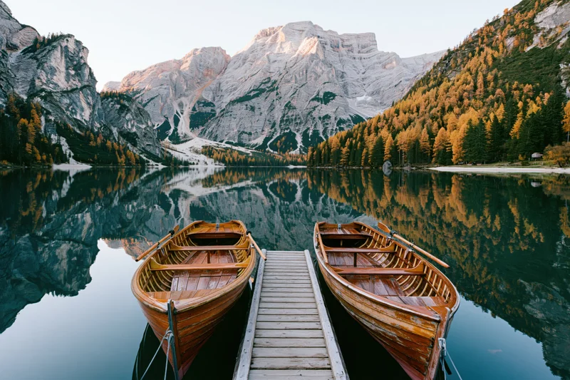 Two wooden rowboats moored beside a small timber dock on a calm alpine lake surrounded by mountains and autumn forest