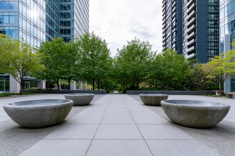 Stone planter bowls arranged along a paved urban courtyard flanked by modern glass buildings and green trees