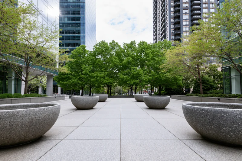 Stone planter bowls arranged in rows along a paved urban courtyard lined with trees and modern buildings