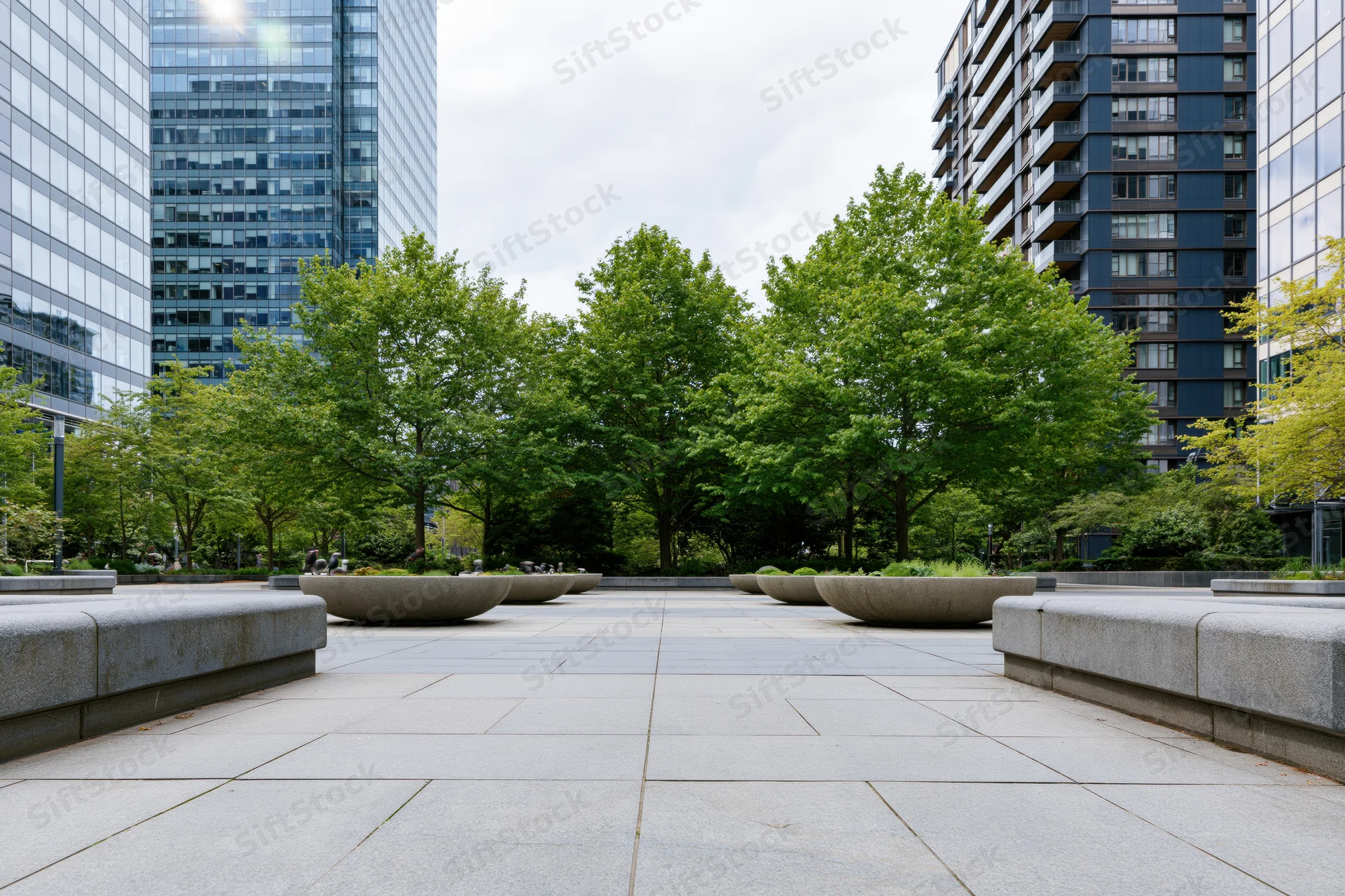 Stone planter bowls lined along a wide urban plaza courtyard with trees and modern buildings surrounding it