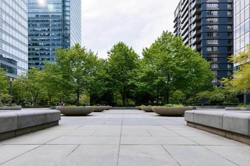 Stone planter bowls lined along a wide urban plaza courtyard with trees and modern buildings surrounding it