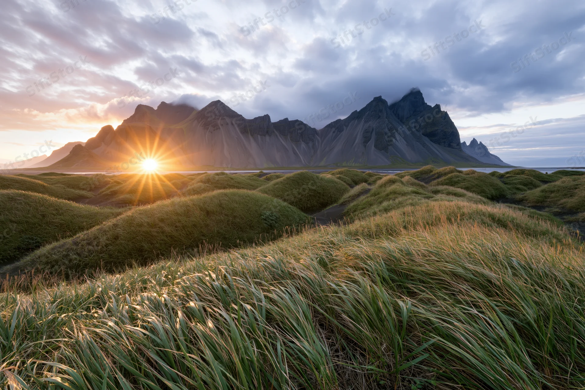 Vestrahorn mountain range rising behind rolling grass-covered dunes at sunset with sunburst over the horizon