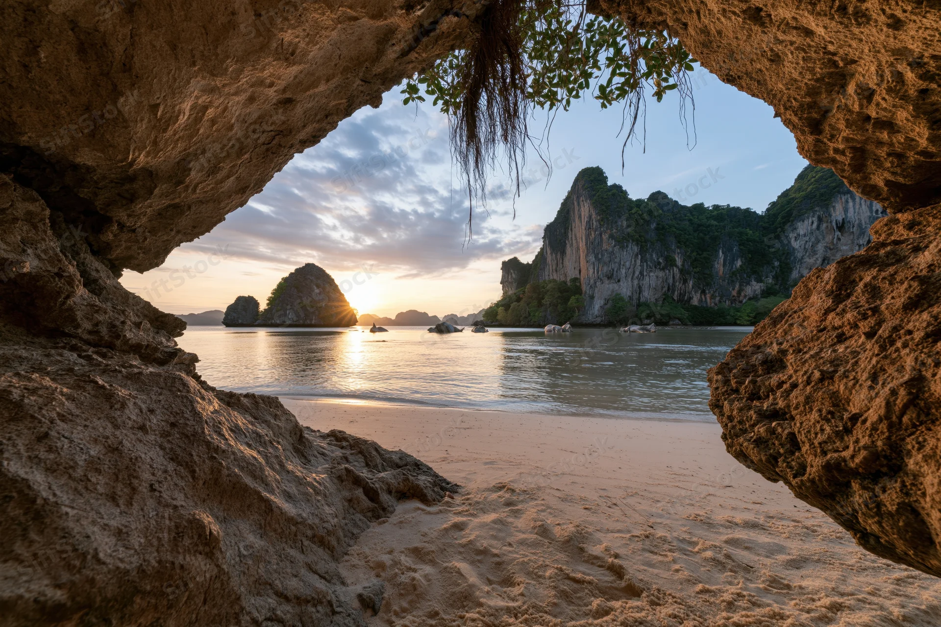 View from inside a shallow sea cave looking out over a calm tropical beach at sunset with karst cliffs and islands in the distance