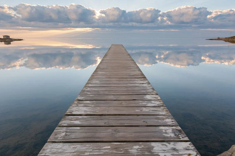 Weathered wooden dock extending into calm bay at dawn with cloud reflections on water surface
