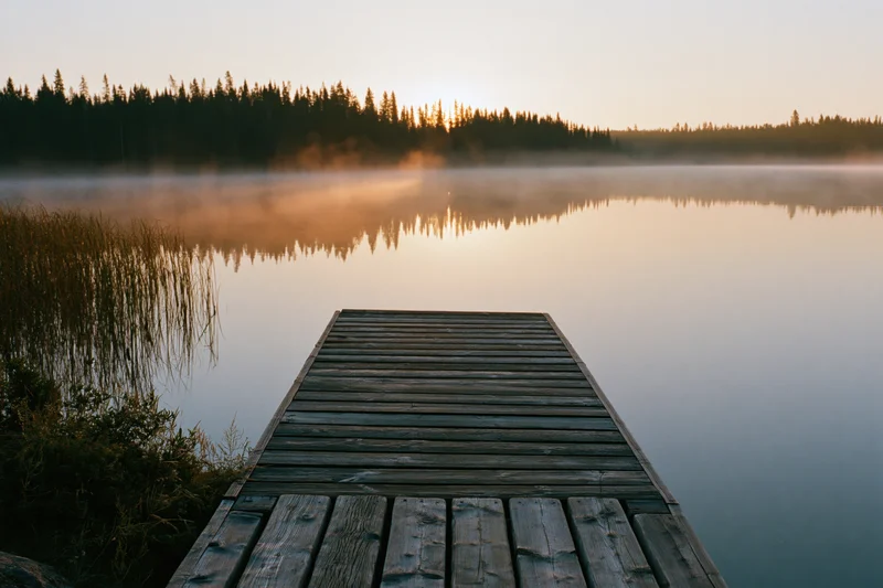 Weathered wooden dock extending over calm misty lake at sunrise with forest reflection in water