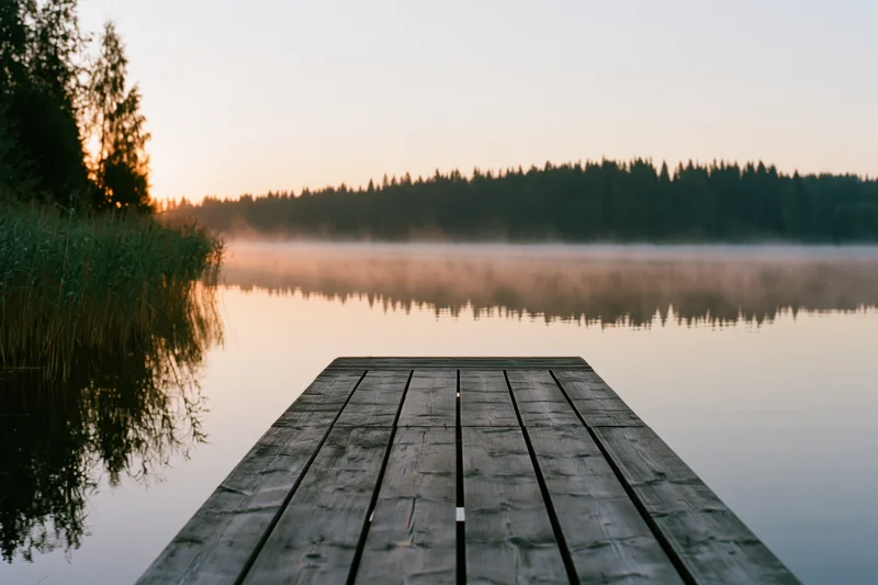 Weathered wooden dock extending over calm misty lake at sunrise with forested shoreline in background