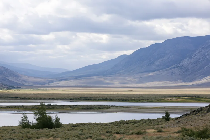 Wide view of a mountain range rising above a river valley with grasslands and shrubs under an overcast sky
