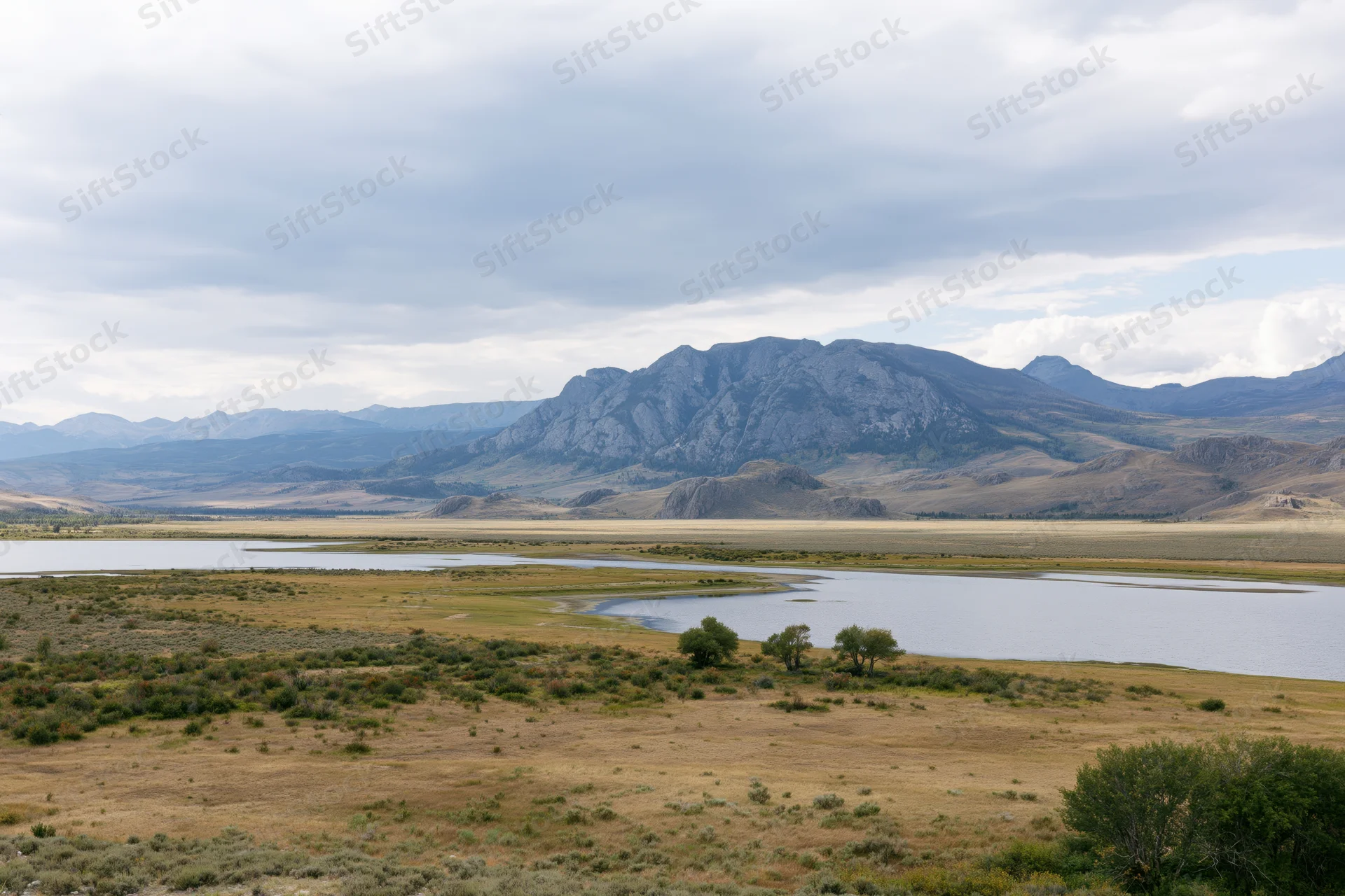 Rocky mountain range rising above a wide valley with a winding river and grassy plains under an overcast sky