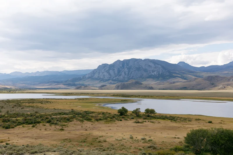 Rocky mountain range rising above a wide valley with a winding river and grassy plains under an overcast sky