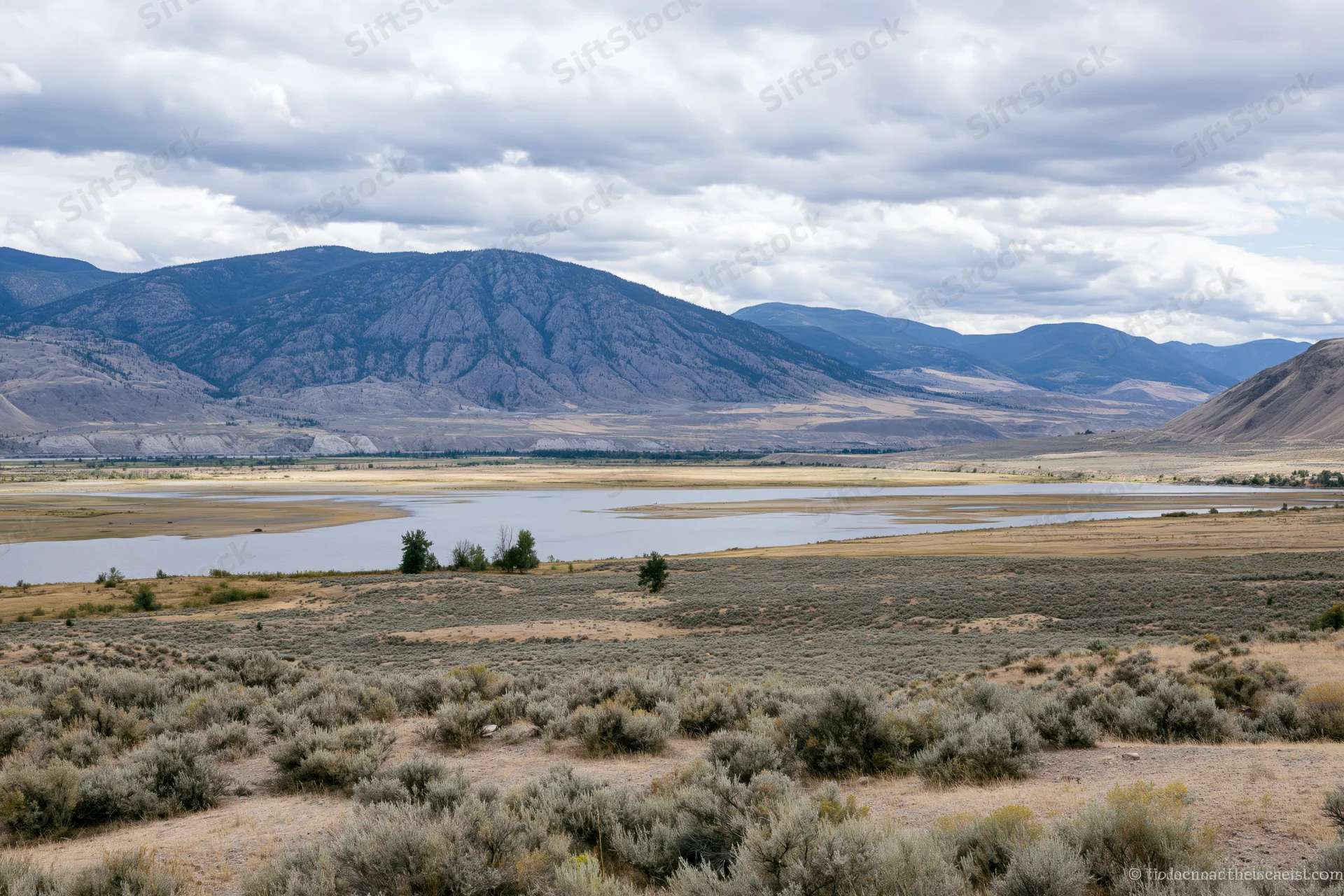 Wide view of a river winding through a sagebrush valley with rocky mountains in the background under cloudy sky