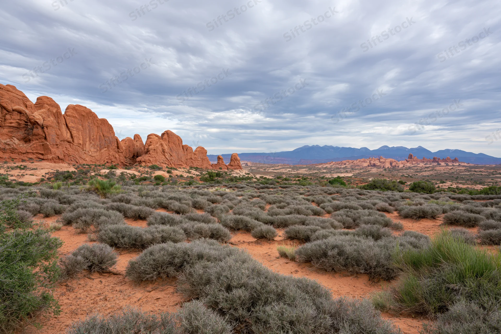 Red sandstone rock formations rising from desert landscape with shrubs and distant mountains under cloudy sky
