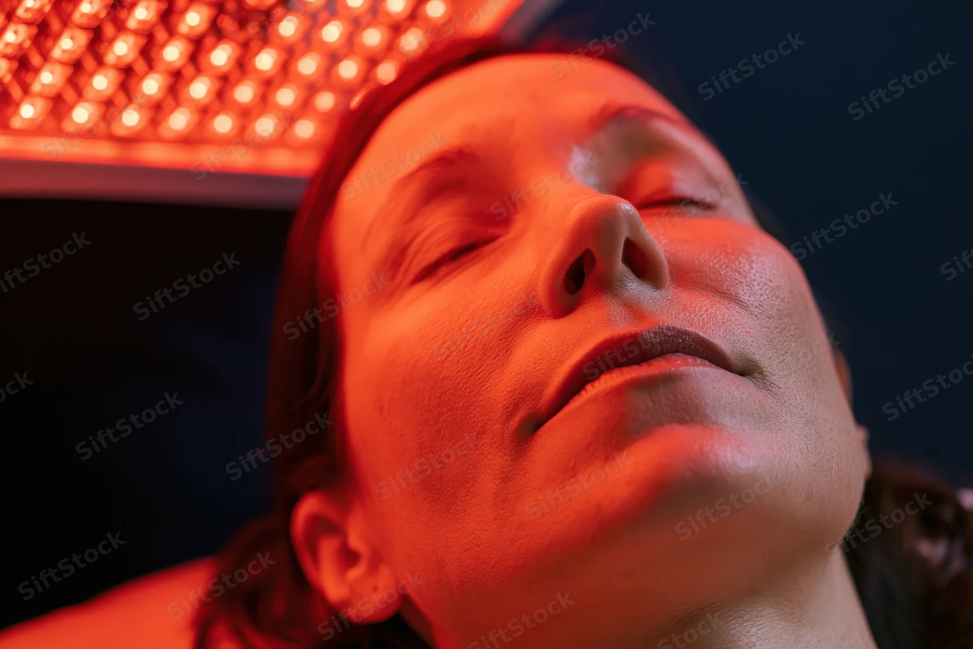 Woman lying back with eyes closed under red LED light for facial therapy