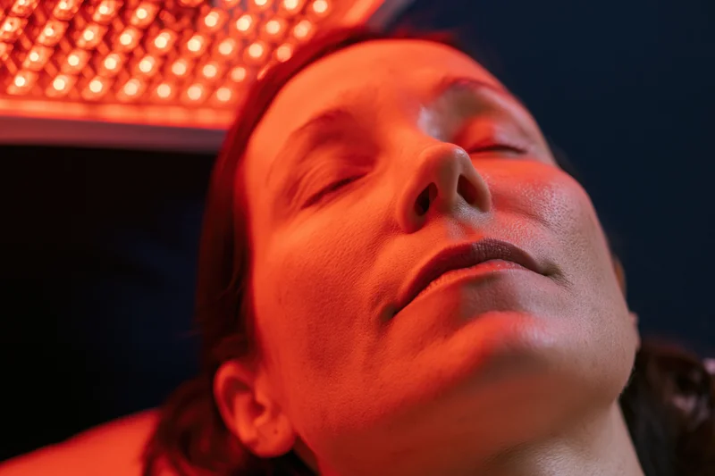 Woman lying back with eyes closed under red LED light for facial therapy