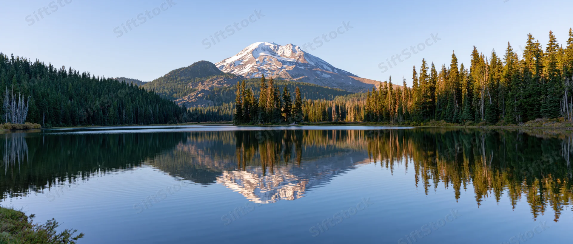 Snow-capped mountain reflected in calm lake surrounded by evergreen forest