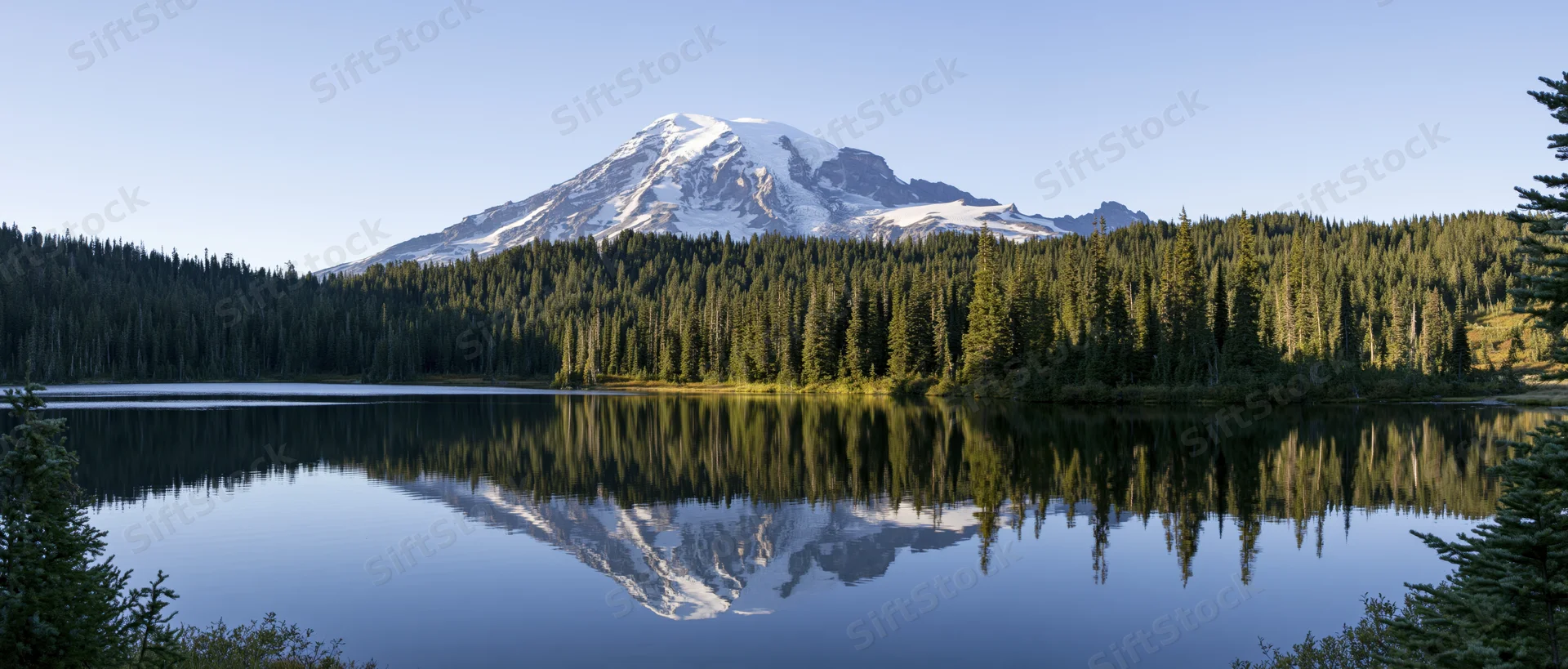 Snow-capped mountain reflected in calm lake surrounded by dense evergreen forest