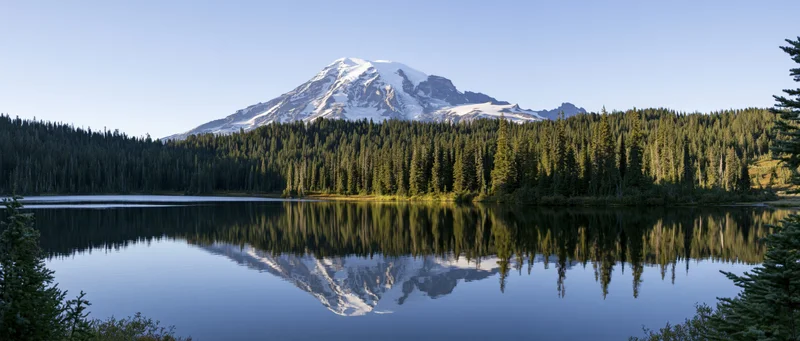 Snow-capped mountain reflected in calm lake surrounded by dense evergreen forest