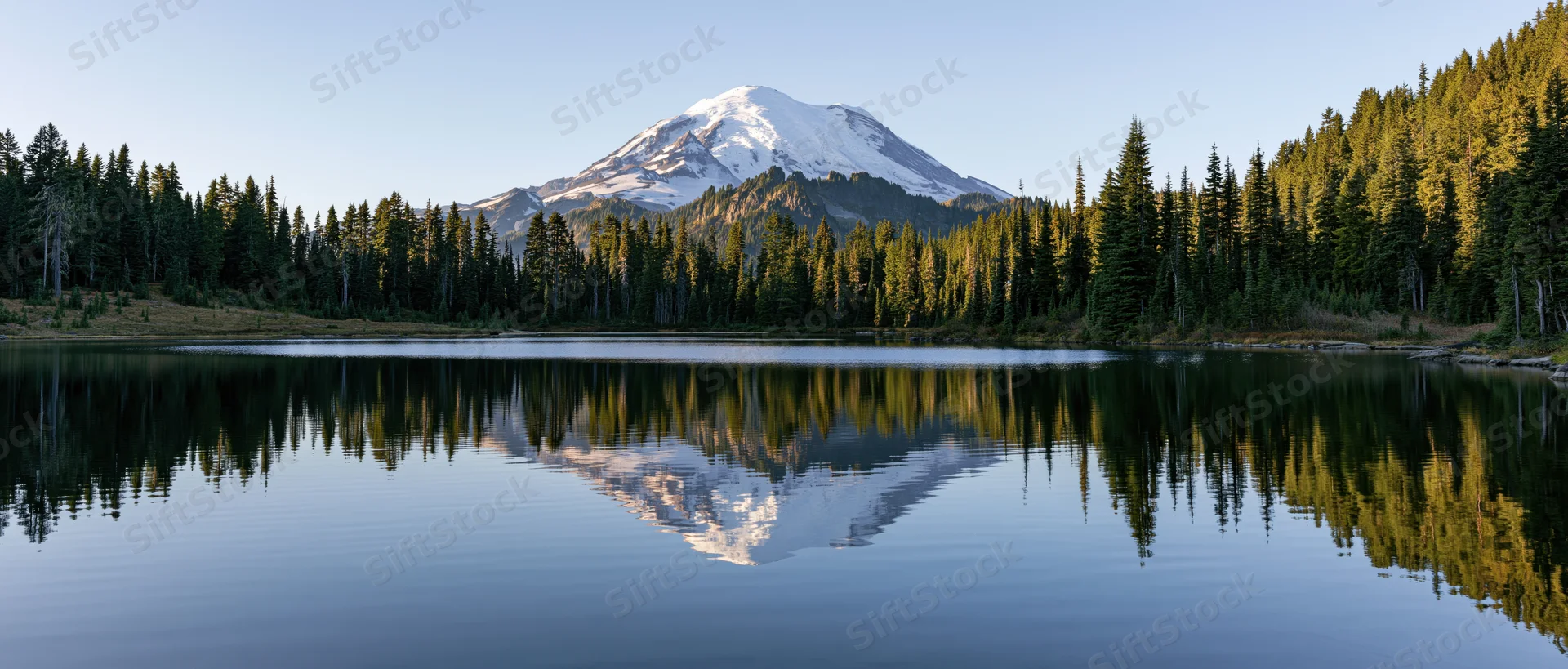 Snow-capped mountain reflected in calm lake surrounded by evergreen forest