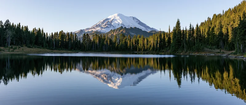 Snow-capped mountain reflected in calm lake surrounded by evergreen forest
