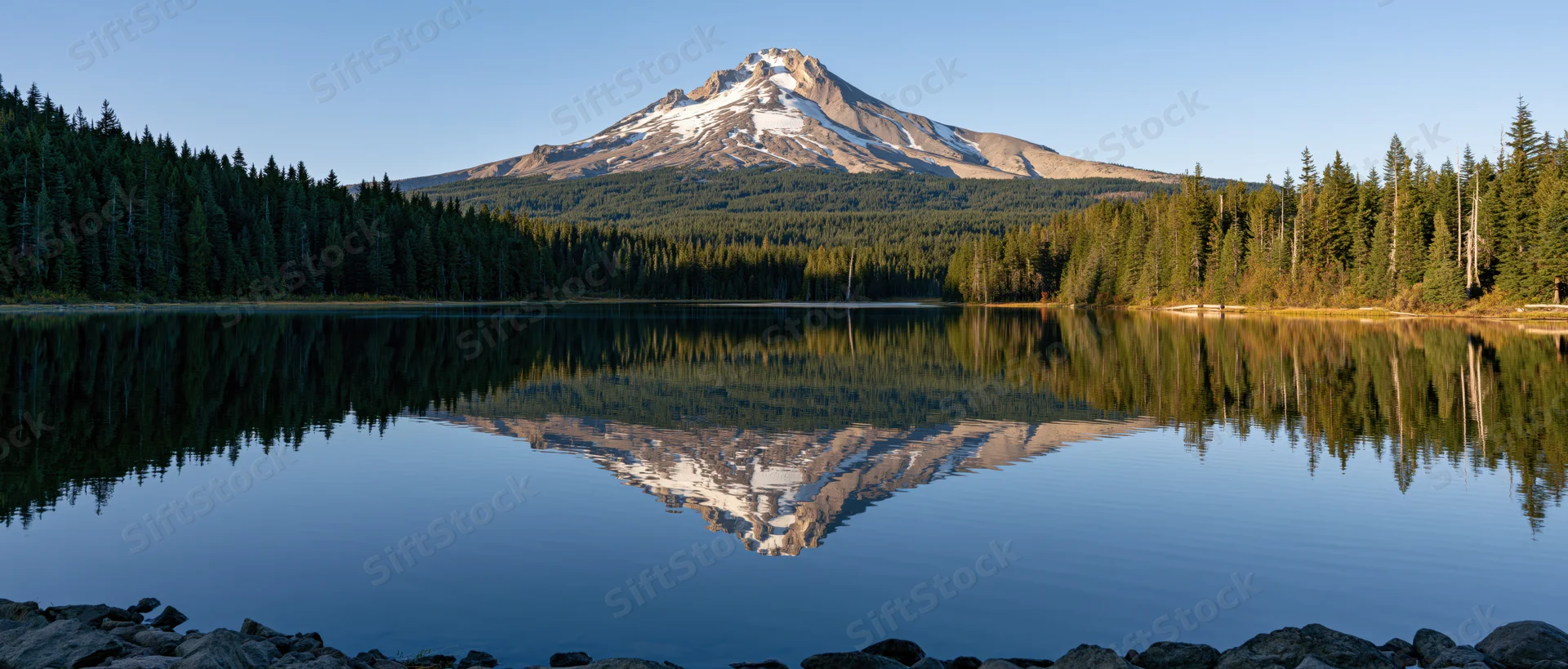 Snow-capped mountain reflected in calm lake surrounded by evergreen forest