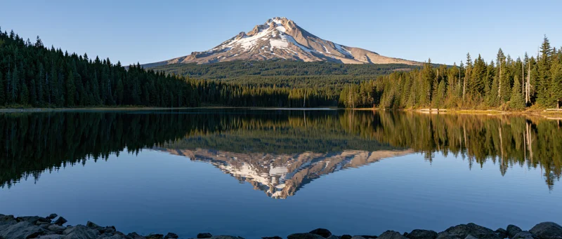 Snow-capped mountain reflected in calm lake surrounded by evergreen forest