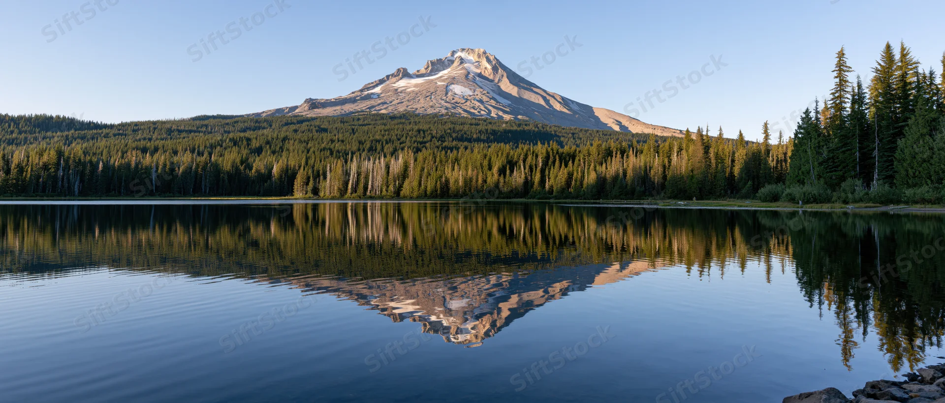 Snow-capped mountain reflected in calm lake surrounded by dense evergreen forest