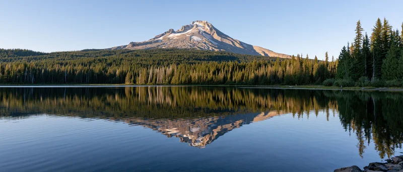 Snow-capped mountain reflected in calm lake surrounded by dense evergreen forest
