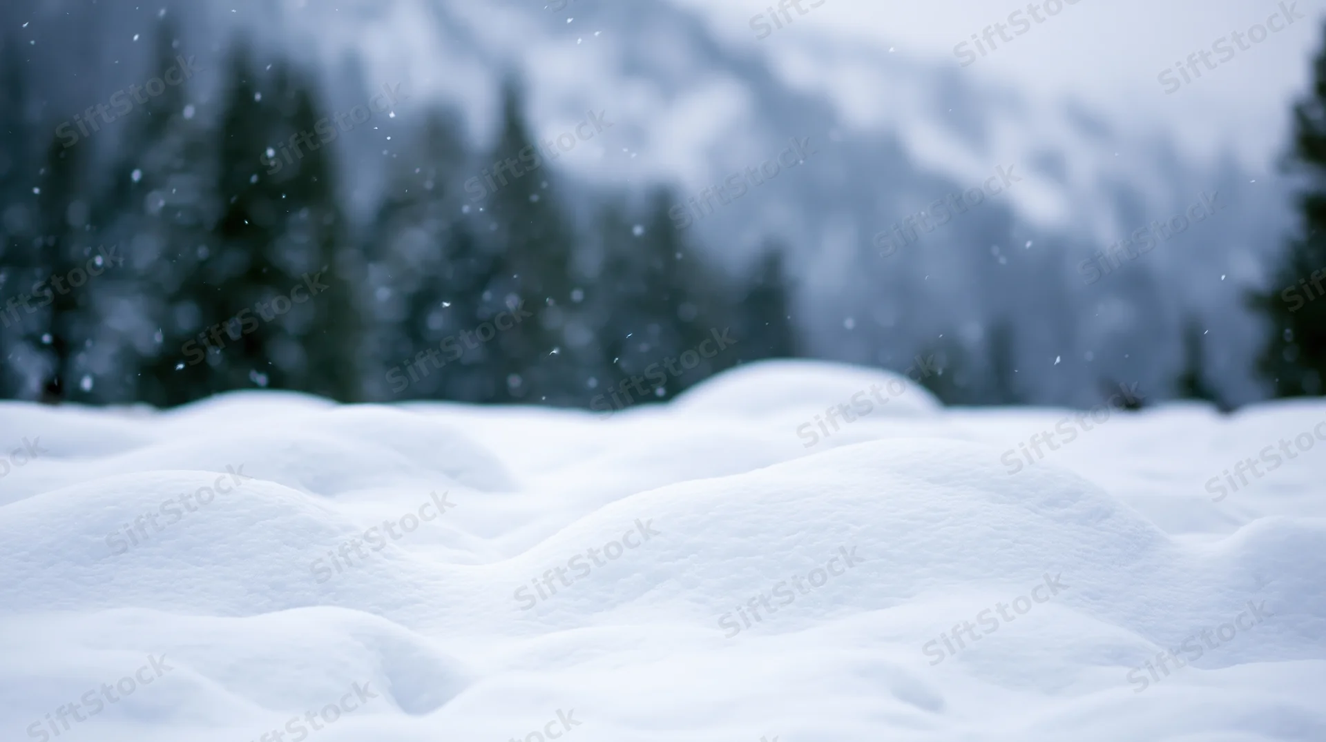 Snow-covered ground with drifting snow in front of blurred evergreen forest and mountains