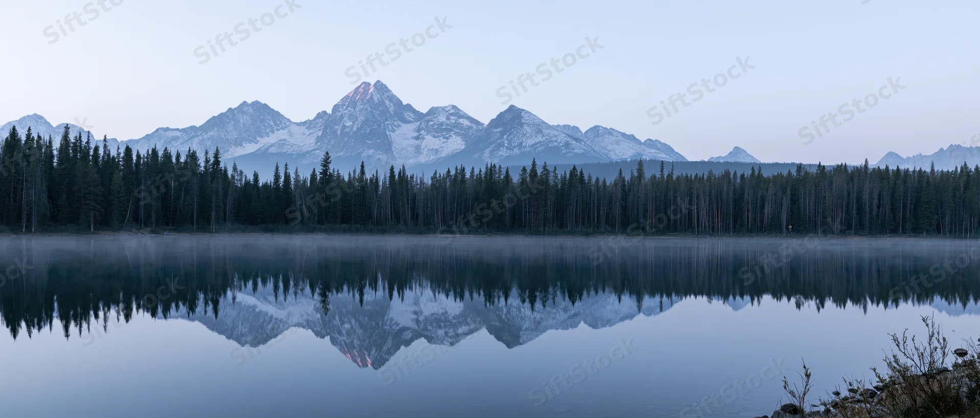 Snow-capped mountain range reflected in calm lake at dawn surrounded by evergreen forest