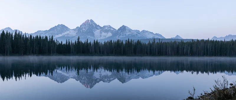 Snow-capped mountain range reflected in calm lake at dawn surrounded by evergreen forest