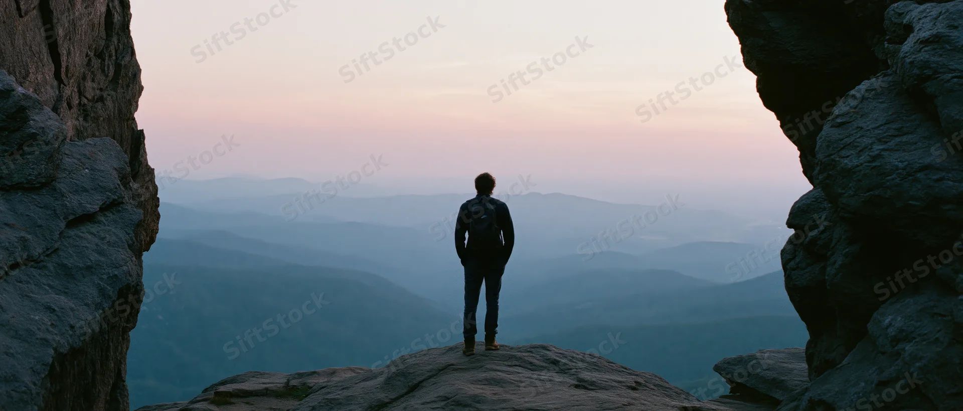 Man with backpack standing on rocky summit overlooking layered mountain valley at dusk