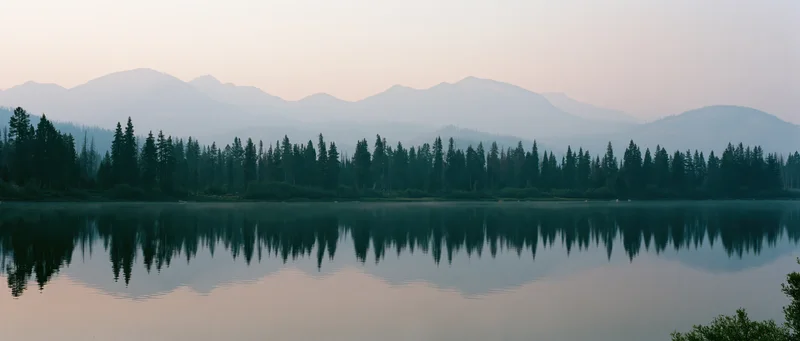 Calm lake reflecting pine forest and mountains at dusk