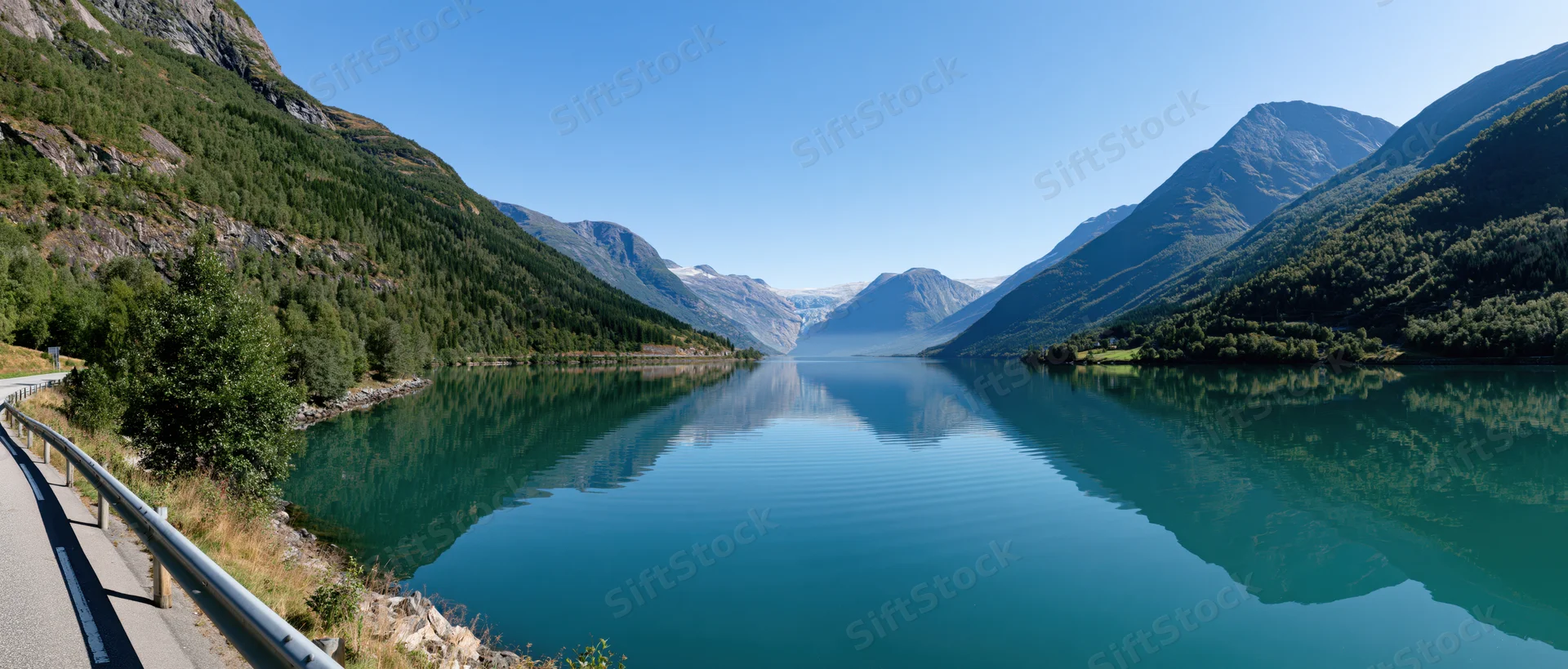 Calm turquoise fjord reflecting mountains and forest under clear blue sky beside winding road with guardrail