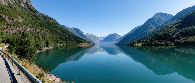 Calm turquoise fjord reflecting mountains and forest under clear blue sky beside winding road with guardrail