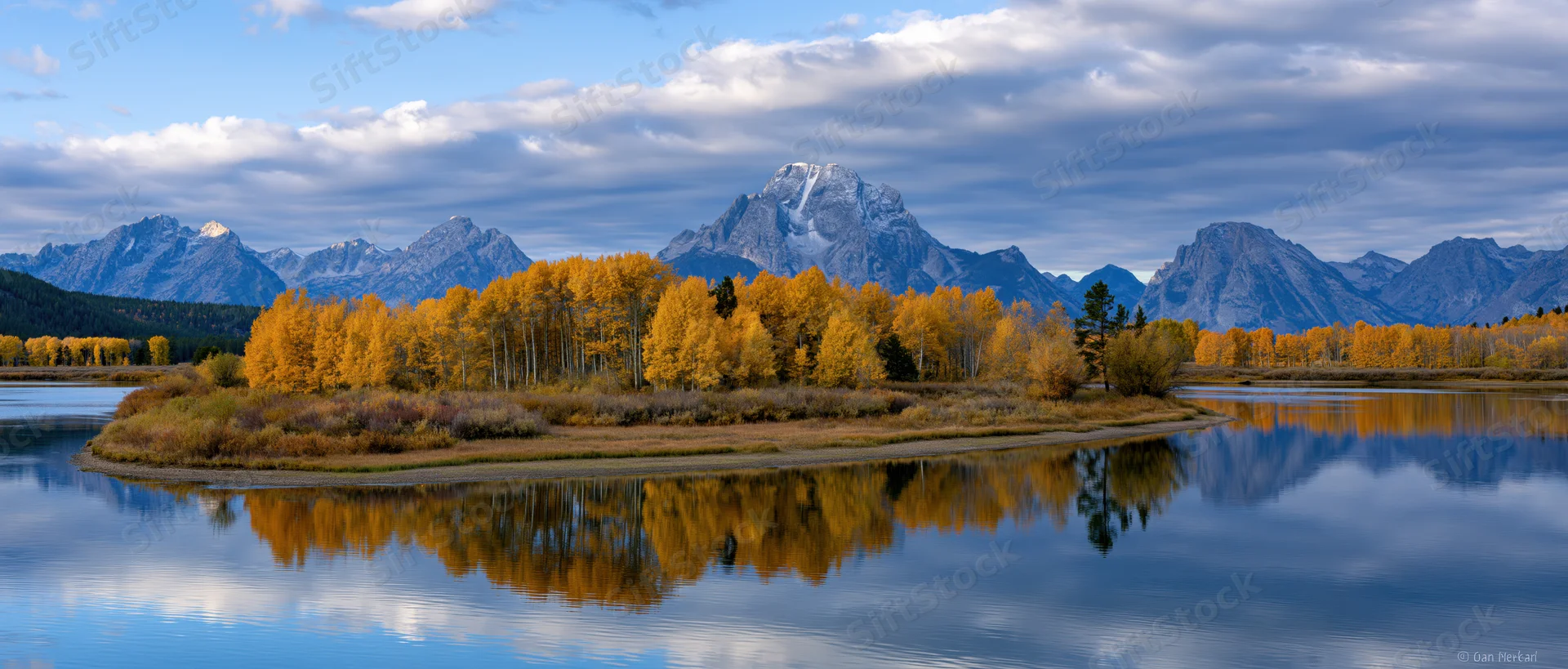 Golden aspen trees lining a river island reflecting in calm water with snow-capped mountains in the background