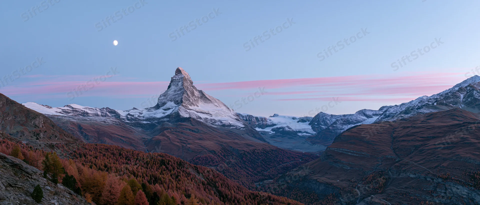 Matterhorn mountain peak rising above autumn forested valley at twilight with moon in sky