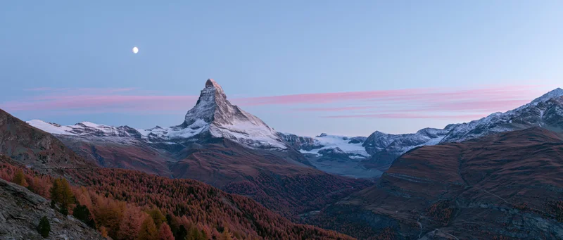 Matterhorn mountain peak rising above autumn forested valley at twilight with moon in sky
