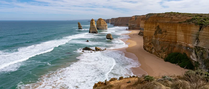 Sea stacks and cliffs along a sandy beach with waves crashing under a blue sky