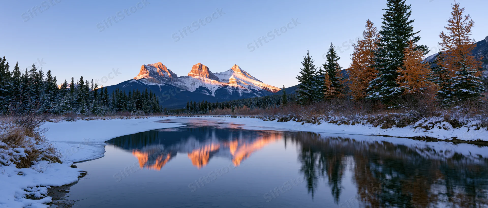 Snow-covered mountains reflected in a calm river surrounded by evergreen and larch trees in a winter forest landscape at sunrise