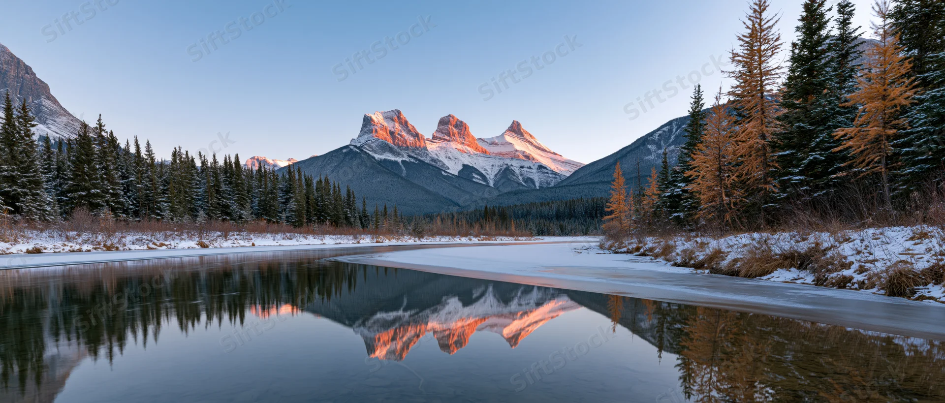 Snow-covered mountain peaks reflected in a still river surrounded by evergreen forest at sunrise