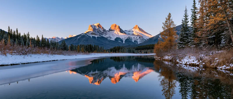 Snow-covered mountains reflecting in a calm river at sunrise surrounded by evergreen forest