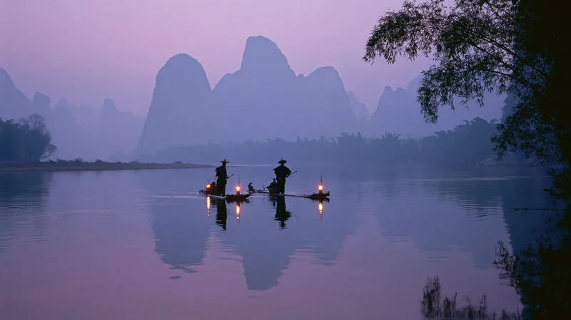 Fishermen with cormorants and lanterns on bamboo rafts at twilight on a calm river with karst mountains in the background
