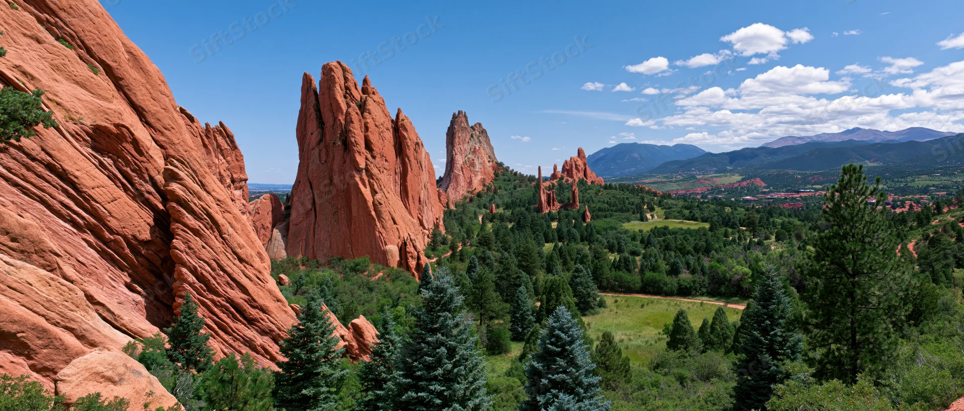 Red rock formations rising from a green valley with pine trees under a blue sky